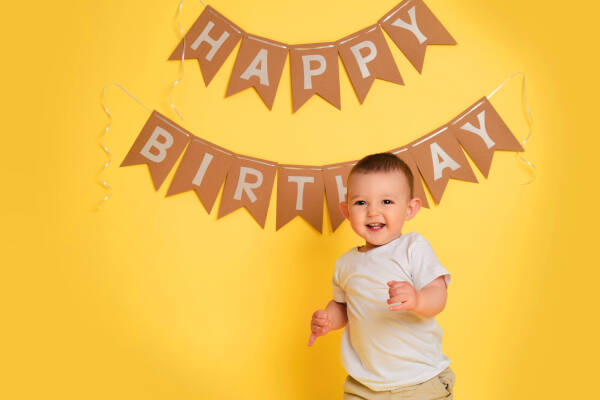 A one-year-old boy stands in front of a yellow backdrop, smiling, with a "Happy Birthday" banner hanging on the wall behind him.