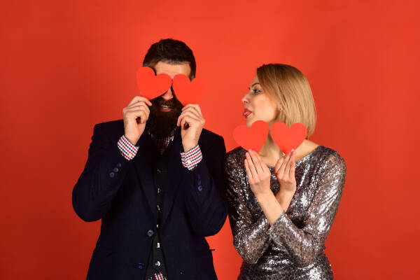 A playful couple poses with red paper hearts, the man covering his eyes and the woman playfully sticking out her tongue in front of a red background.