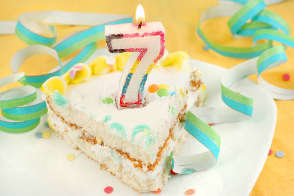 A slice of cake with a lit number 7 candle, surrounded by colorful confetti and party streamers on a white plate.