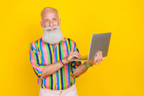 A smiling elderly man with a long white beard holds a laptop, wearing a colorful striped shirt against a yellow background.