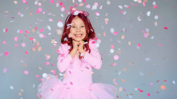 A smiling young girl in a pink tutu dress, surrounded by colorful confetti falling in the air against a gray background.