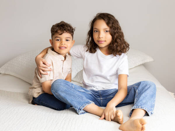 A young boy and girl sit together on a bed, smiling at the camera, with the older sibling's arm around the younger one.