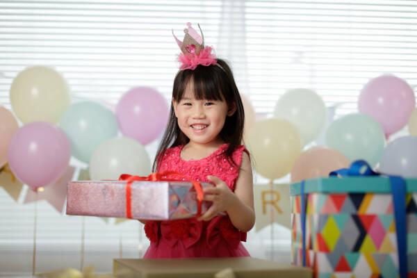 A young girl in a pink dress and crown smiles while holding a wrapped gift, surrounded by colorful balloons and presents.