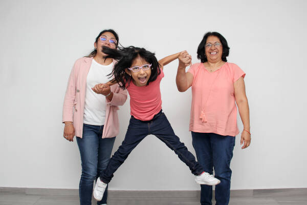 A young girl joyfully jumps while holding hands with two women, all of them smiling and wearing casual outfits.