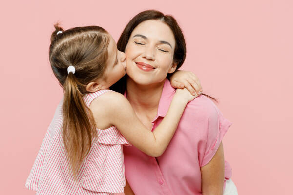 A young girl kisses her mother on the cheek, both wearing pink, with a light pink background behind them.