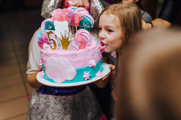 A young girl leans in to take a bite of a colorful birthday cake decorated with pink and blue frosting and a number 5 topper.