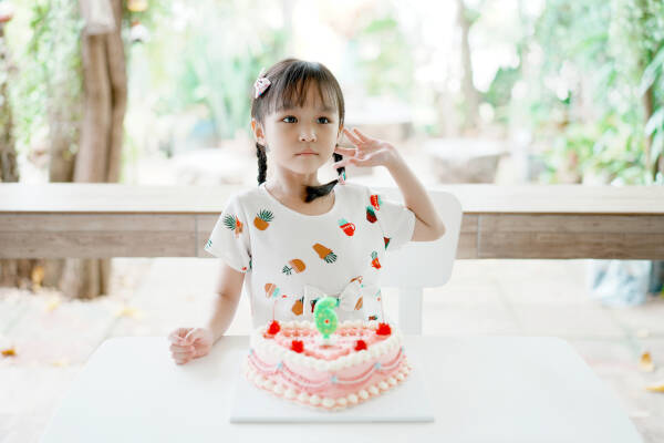 A young girl sits at a table with a decorated birthday cake in front of her, holding her pigtail and looking thoughtful.