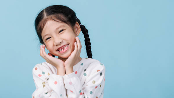 A young girl with braided hair, smiling brightly with her hands on her cheeks, wearing a polka-dotted sweater against a blue backdrop.