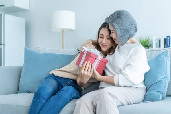 A young woman receives a gift from an older woman, both smiling and hugging on a sofa, creating a warm moment.