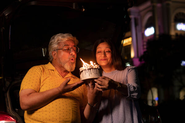 An elderly couple enjoys a late-night birthday celebration outdoors, with the man blowing out candles on a cake.