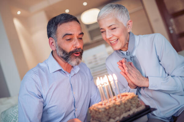 An elderly man and woman are celebrating a birthday, blowing out candles on a cake together, while smiling.