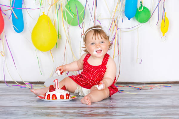 Baby girl celebrates birthday with red cake, in a red dress, with balloons on white backdrop.