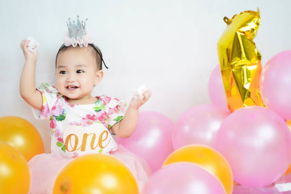 Baby girl, wearing crown, celebrates first birthday amidst colorful balloons on white background.