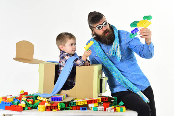 Bearded father plays with his little son, pretending a cardboard box is an airplane, surrounded by Lego.