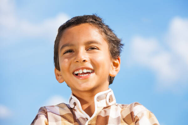 Close-up of a young boy smiling brightly outdoors with a clear blue sky in the background.