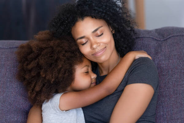 Close-up portrait of a loving mother and her little daughter sitting on the couch at home, hugging with closed eyes.