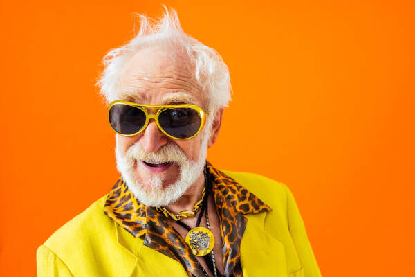 Cool old man flashes a smile for the camera, dressed in bright yellow and leopard print clothes against an orange background