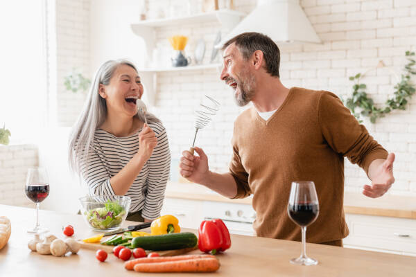 Couple is cooking together, holding utensils as if singing into microphones, surrounded by fresh vegetables in their kitchen.