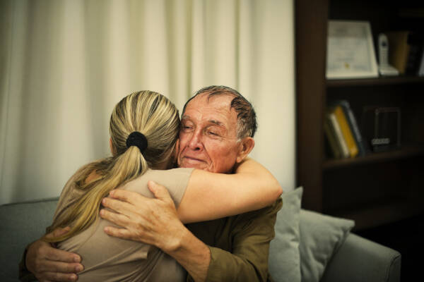 Daughter tenderly embraces her elderly father as he sits on the sofa at home.