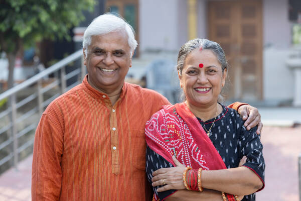  Elderly couple standing together, smiling at the camera while wearing traditional clothing, outdoors in a home setting.