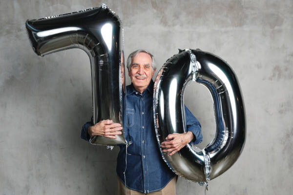 Elderly man holding large silver balloons shaped like the number 70, celebrating his birthday against a gray background.