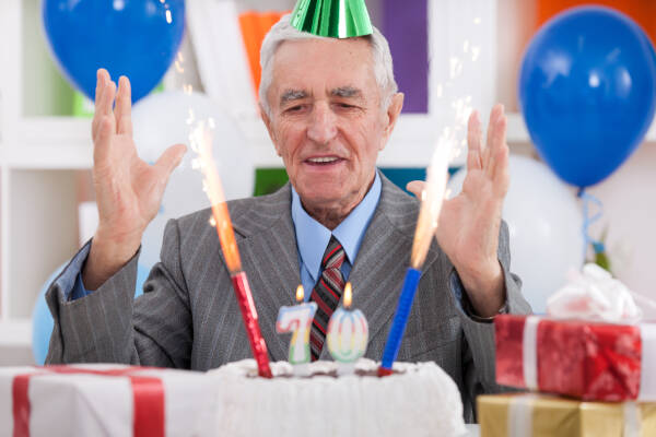 Elderly man in a suit and party hat celebrating his 70th birthday with sparklers on a cake, surrounded by gifts and balloons.