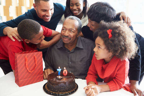 Elderly man sitting at table with a birthday cake and the number 70 on it, surrounded by smiling family members and children.