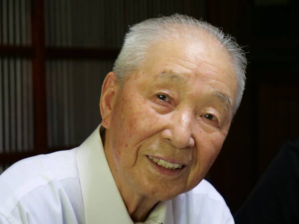 Elderly man smiles warmly into the camera against a dark background.