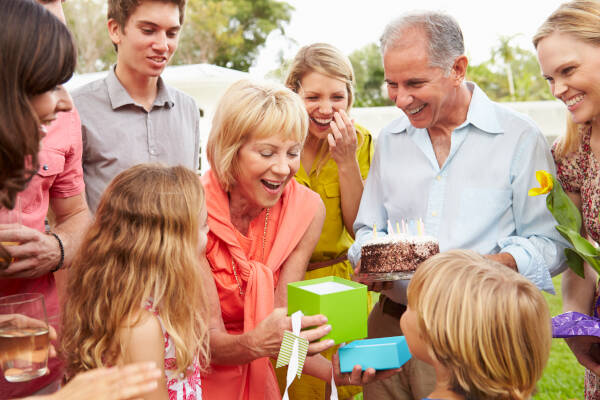 Family gathering outdoors, celebrating with cake and gifts, while an older woman opens a present with excitement.