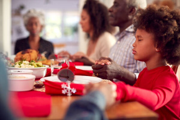 Family sitting at a dinner table holding hands, eyes closed in prayer before a meal during a festive holiday gathering.