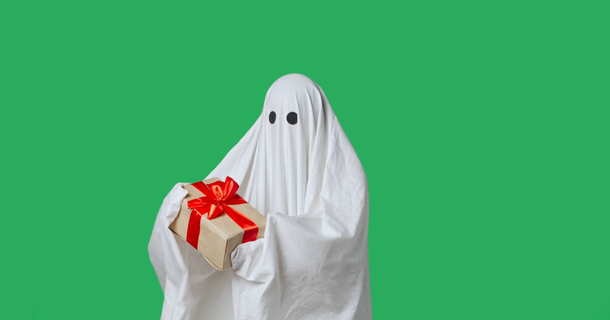 A person dressed as a ghost, covered in a white sheet with black eyes, holds a gift box wrapped with a red ribbon. The background is a solid green.