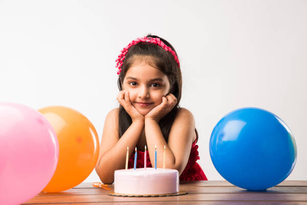 Girl with a headband rests her chin on her hands, smiling in front of a birthday cake with lit candles and colorful balloons.