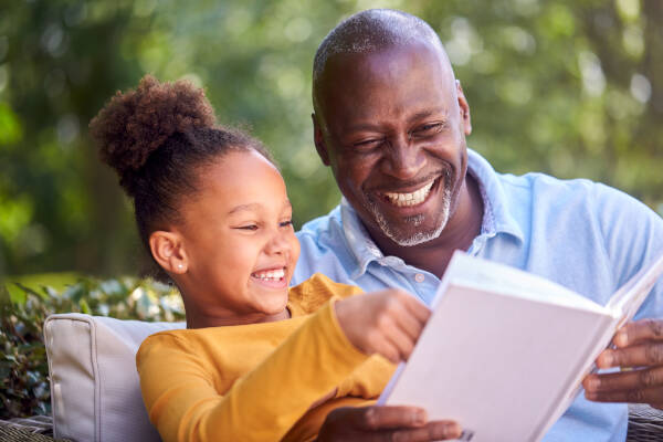 Grandfather and young girl reading a book together, both smiling and enjoying a bonding moment outdoors.