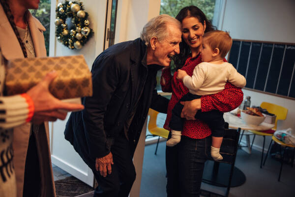 Grandfather joyfully greeting a baby held by his mother as family members gather for a holiday celebration.