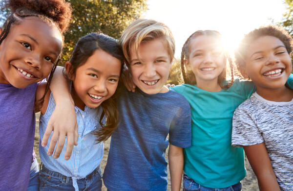 Group of diverse kids standing arm in arm, smiling outdoors on a sunny day, with sunlight filtering through the trees.