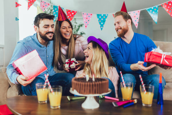 Group of friends smiling and laughing as they pass gifts to the birthday girl, with a cake on the table.