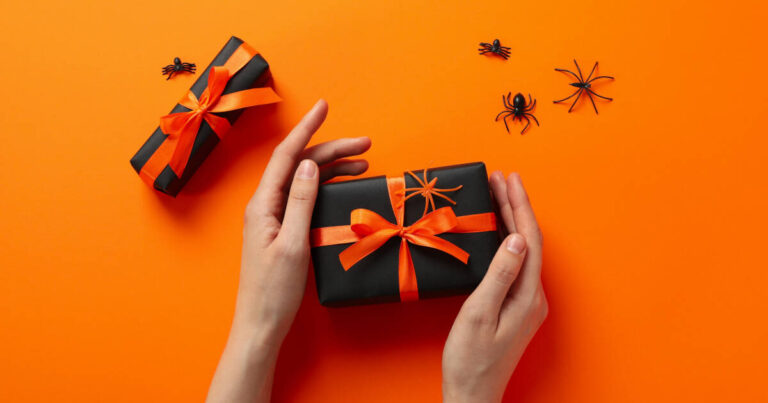 A pair of hands holding a black gift box wrapped with an orange ribbon, placed against an orange background. Small black plastic spiders are scattered around.