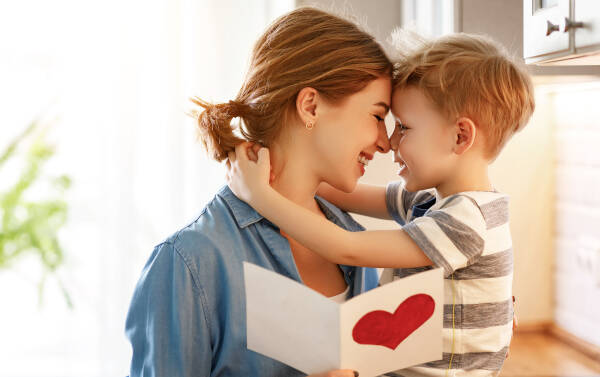 Happy little boy congratulates smiling mother, presenting a card with a red heart during Mother's Day celebration at home.