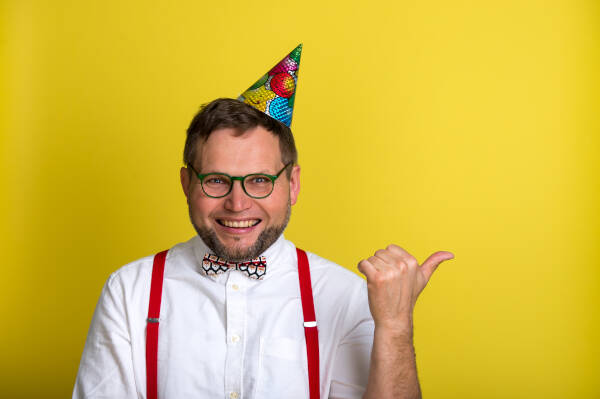 Nerdy man smiles in a party hat and bowtie, pointing with thumb on a yellow backdrop.