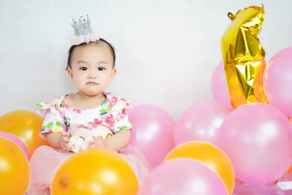 One-year-old girl sits surrounded by a sea of gold and pink balloons, wearing a dress and tiara, against a white background.