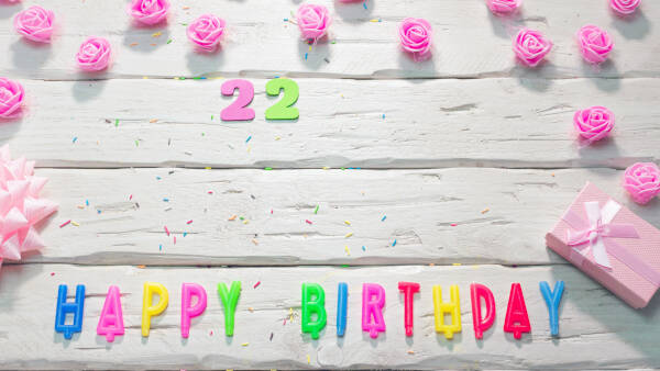 Paper '22' above 'HAPPY BIRTHDAY' candles, set against a wooden backdrop with pink flowers and gift boxes