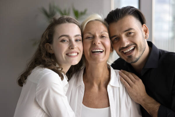 Siblings hugging their senior mother with their cheeks touching, all smiling, as they stand in someone's home.