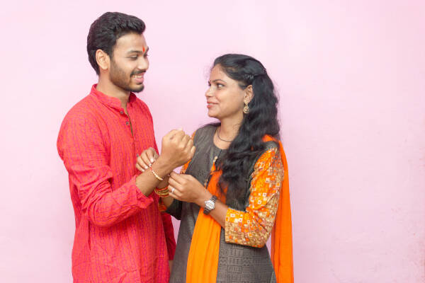 Sister ties a bracelet around her brother's wrist as they both talk and smile against a pink backdrop.