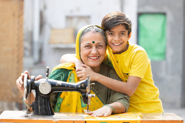 Smiling boy hugging his grandmother while she works on a sewing machine outside, both wearing matching yellow clothes.