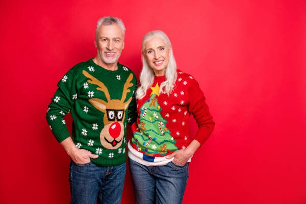 Smiling older couple wearing festive Christmas sweaters, posing together against a red background.