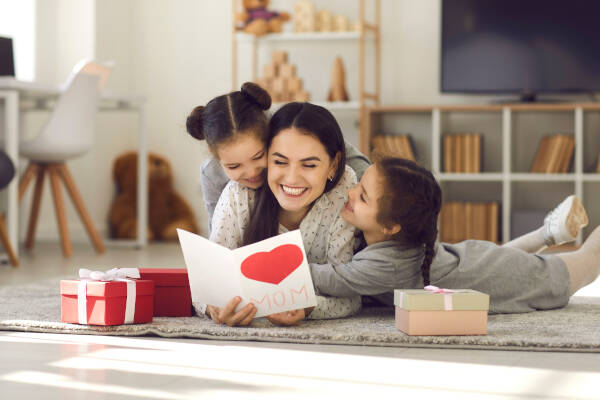 Smiling woman receives presents from kids. Twin daughters give handmade cards, hugging mom, wishing Happy Mother's Day.