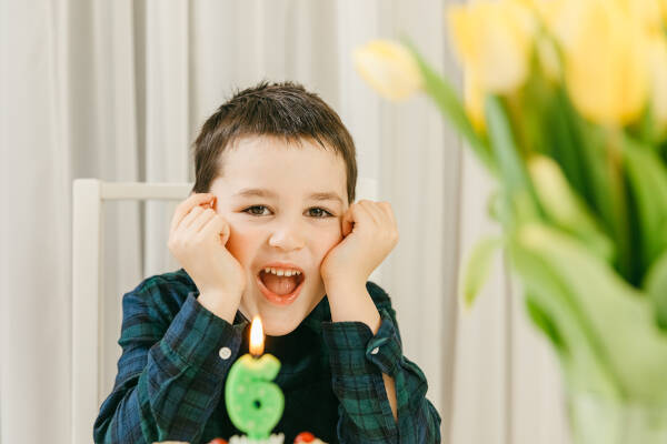 Smiling young boy celebrating his sixth birthday, sitting in front of a birthday cake with a number six candle.