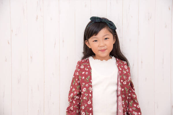 Smiling young girl with a black bow in her hair, standing against a light wooden background.