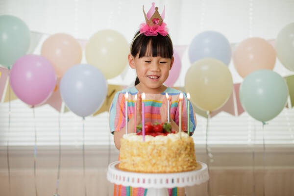 Smiling young girl with a pink crown on her head, celebrating her birthday in front of a cake with lit candles.