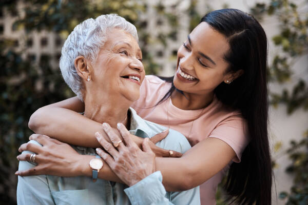 The 32-year-old daughter hugs her elderly mother from behind as they both smile at each other.